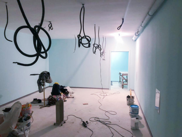 Middle stage of reconstruction in former bomb shelter showing finished walls and partially completed new wiring hanging down from ceiling and construction tools and paint cans sitting on the subfloor.