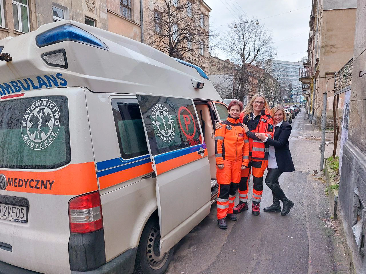 Two people in orange EMT suits and one person in black jacket, white shirt and black leggings stand beside open side door of ambulance on a city street.