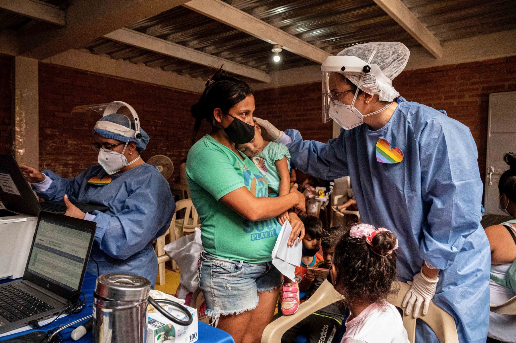 Doctor in surgical gown with rainbow pin on front and wearing mask treats mother holding baby. Another child sits in chair in front and another health care provider works on a computer beside the doctor and mother.