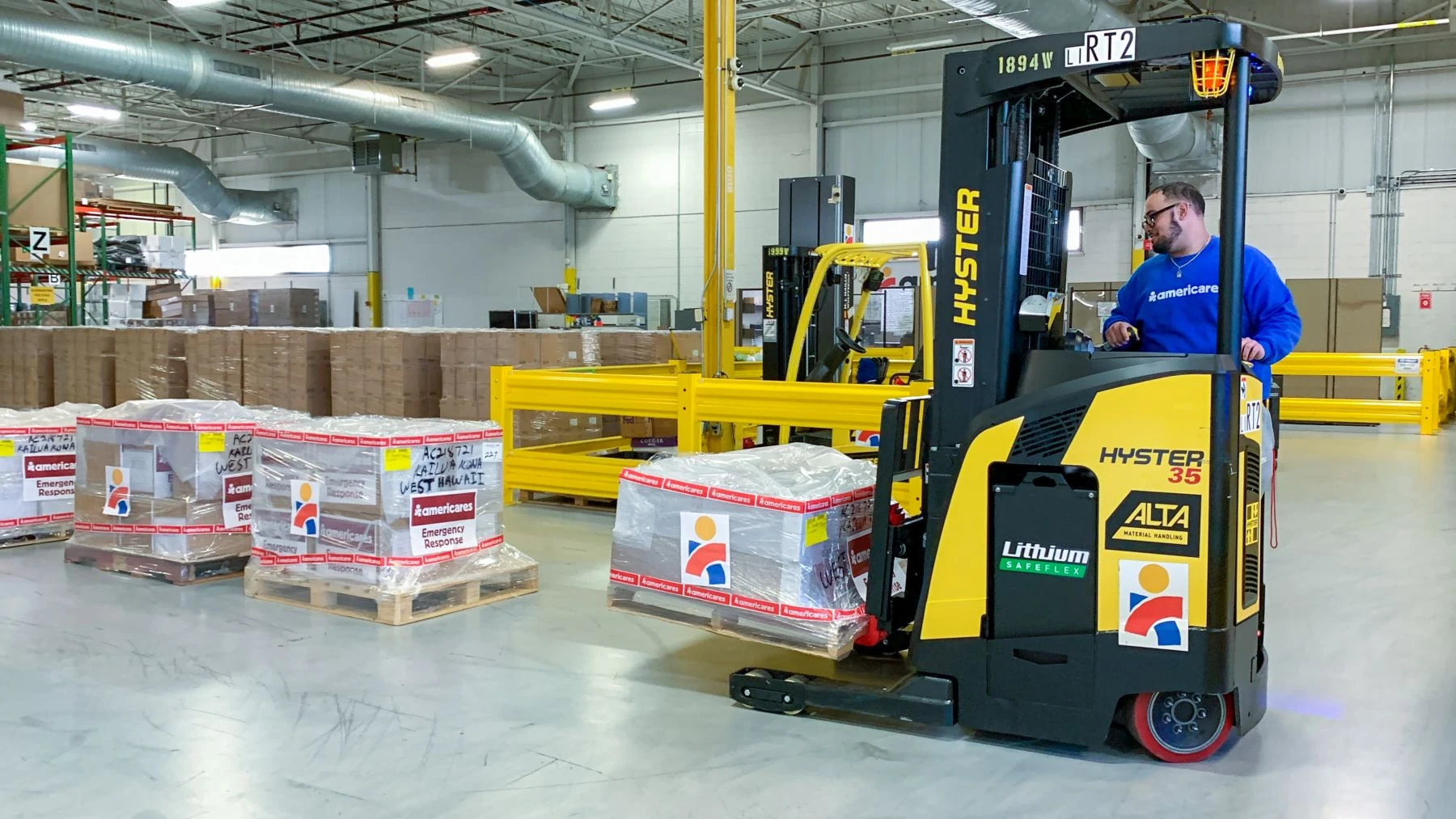 Staff member in blue sweatshirt operating forklift to ready relief supply shipment for Hawaii. Four palettes of supplies are set in a row.