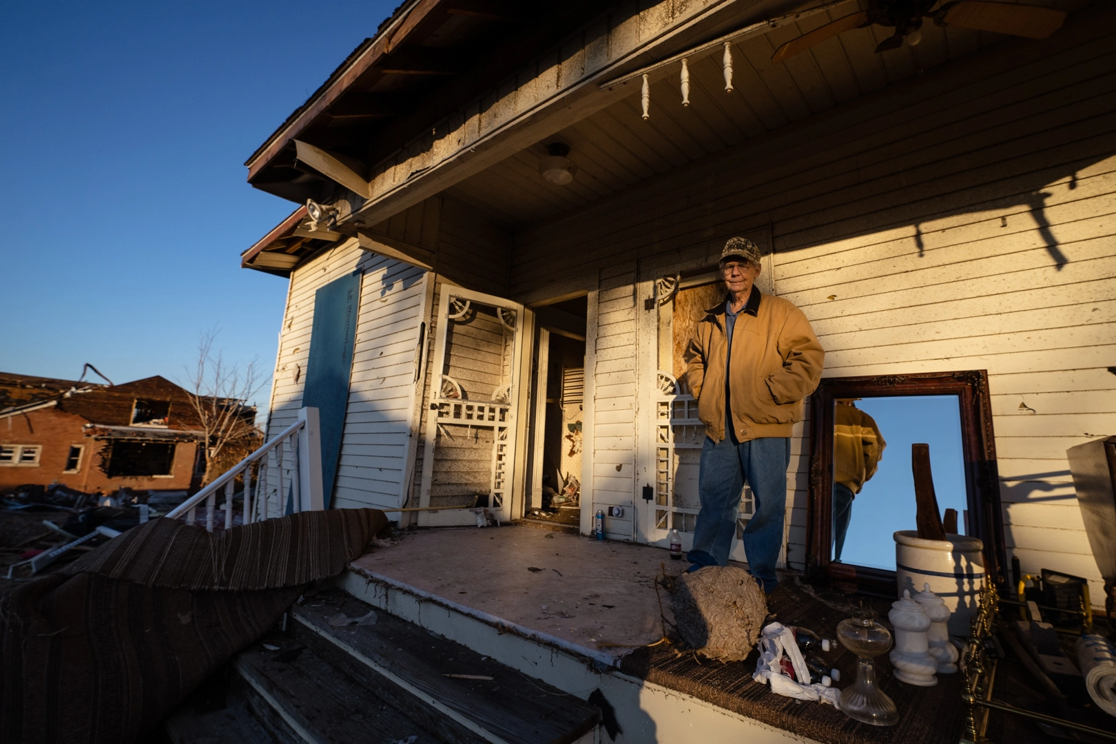 Individual stands on the remains of the porch of a ruined house next to a few salvaged possessions