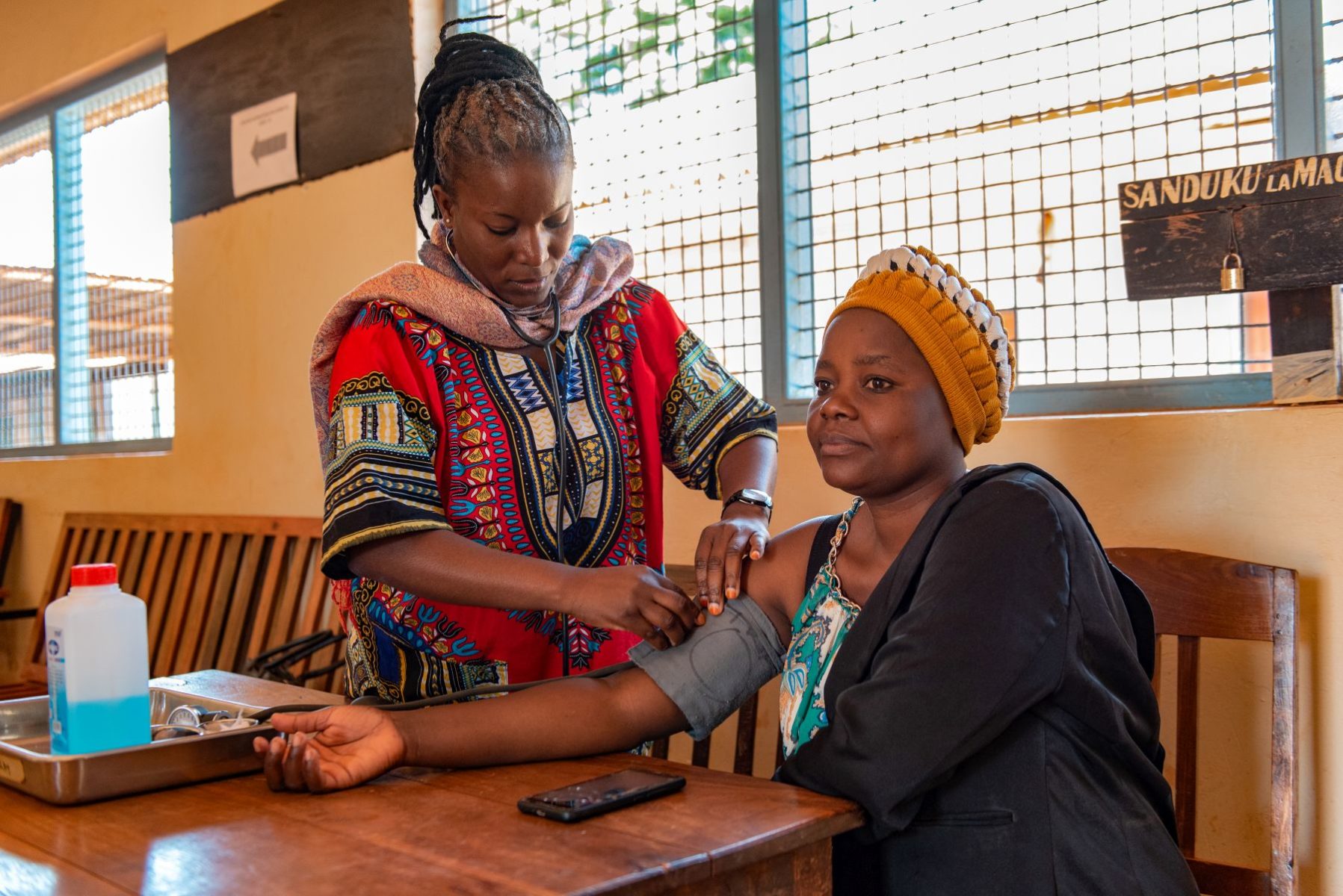 Patient on right in yellow knit cap has her blood pressure taken by nurse in red and multi-patterned dress with stethoscope. Tray on table with medical supplies. Window to outside in background.