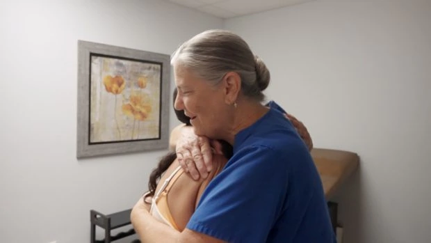 Health worker in blue embraces patient in exam room.