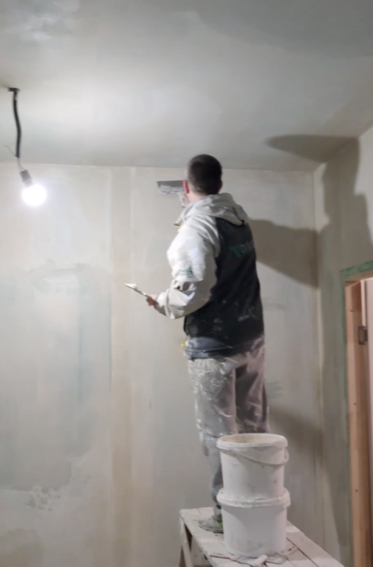 Workman is working on the sheet rock for the new cancer storage facility. He stands on a bench with buckets of spackle behind him while he works on the wall with back to us.