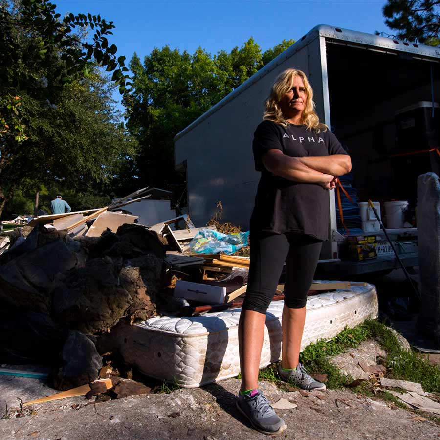 Woman looks in the distance with crossed arms with a mattress, destroyed home contents, and a moving truck behind her.