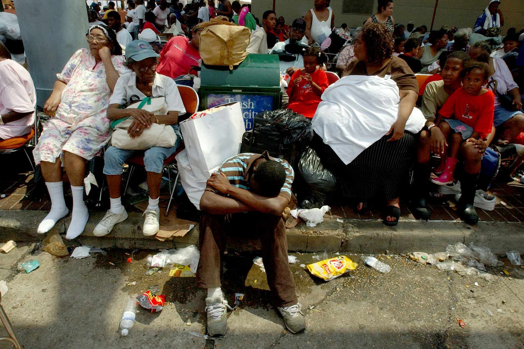 A crowd of refugees sit with plastic bags of their belongings on the curb of a street.